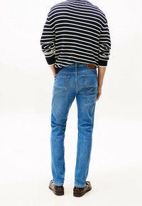 Persona de pie con las manos en los bolsillos, vistiendo jeans azules, zapatos marrones y un suéter a rayas en azul marino y blanco, de espaldas frente a un fondo liso.