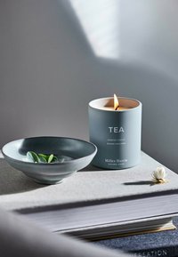 Duftkerze in einem hellblauen Behälter, beschriftet mit "TEE", mit angezündetem Docht. Begleitet von einer dunkelgrauen Schale mit grünen Blättern, auf strukturierten Oberflächen.