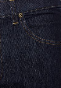 Close-up van een donkerblauwe spijkerbroekkzak met oranje stiksels en een kleine metalen klinknagelknop vlak bij de hoek van de zak.