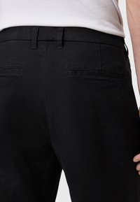 Pantaloni neri con una texture liscia, caratterizzati da una vita piatta, due tasche posteriori e sottili dettagli di cucitura.