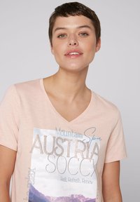 Jasnoróżowa koszulka z dekoltem w serek z graficznym nadrukiem zawierającym tekst i motywy górskie, ozdobiona kryształkami dla dodatkowego efektu.