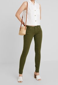 Pantalones ajustados verde oliva, blusa sin mangas blanca de ojo con botones y mules trenzados color blanco roto. Incluye un pequeño bolso redondo de paja.