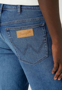 Blue denim jeans with a Wrangler logo patch and distinctive W-shaped stitching on the back pockets. Features a straight leg design.