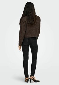 Pruun, tekstuuriga lühike jakk, millel on pikad varrukad, koos kitsaste mustade pükste ja mustade kontsakingadega, vaadatuna tagantpoolt.