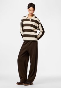 Osoba stojąca z rękami za plecami, ubrana w kremowo-brązową koszulę w paski z długim rękawem, luźne ciemnobrązowe spodnie i brązowe buty.