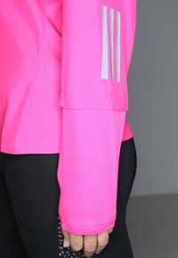 Personne portant un maillot de sport à manches longues rose vif avec des rayures réfléchissantes sur le bras et un legging noir avec des détails à motifs.