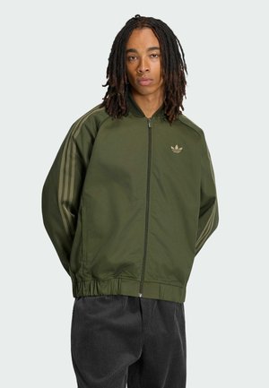 Junger Mann mit langen Dreadlocks, trägt eine grüne Adidas-Jacke mit Reißverschluss und gestreiften Ärmeln sowie dunkle Cordhosen, steht mit den Händen hinter dem Rücken.