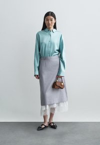 Vaaleansininen nappikauluspaita on yhdistetty harmaisiin hameeseen, jossa on pitsireunus. Mallilla on käsissään ruskea käsilaukku, jossa on kultainen yksityiskohta.