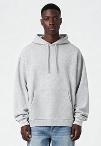 Šedá mikina s kapucňou vyrobená z mäkkej, textúrovanej tkaniny. Obsahuje klokaní vrecko a ribbené manžety a lem. Uvoľnený, nadmerný strih.