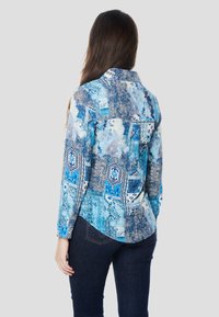 Modrá košeľa s dlhými rukávmi v odtieňoch modrej s abstraktnými a geometrickými vzormi, so zapínaním na gombíky pri golieri a zaobleným lemom. Ľahká tkanina.