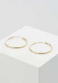 Pendientes de aro en tono dorado con una superficie lisa y brillante y forma circular, exhibidos en un pedestal blanco.