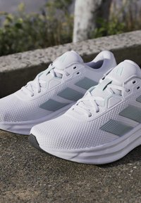 Paire de baskets blanches avec des accents gris clair reposant sur un trottoir extérieur près d'un bord en béton et de la verdure.
