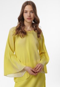 Gele blouse met uitlopende mouwen, gemaakt van lichtgewicht stof met verticale strepen. Rond de halslijn en gelaagde onderrand.