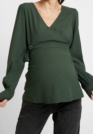 Blusa verde texturizada con diseño cruzado en la parte superior y mangas largas, con escote en V y detalle de lazo lateral, combinada con vaqueros oscuros.