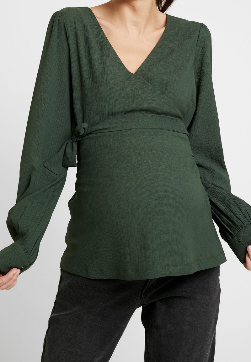 Blouse verte texturée à encolure drapée avec manches longues, présentant un col en V et un détail noué sur le côté, associée à un jean foncé.