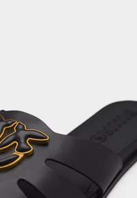 Sandale noire à glissière avec un emblème en forme d'oiseau noir et or sur la bride, et une semelle intérieure texturée avec un logo embossé.