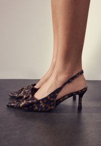 Piedi in décolleté slingback con stampa leopardata marrone e nera, a punta e con tacco kitten basso, su un pavimento scuro.