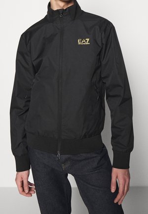 Mężczyzna w czarnej kurtce na suwak z logo "EA7 Emporio Armani" w złotym kolorze na lewej piersi, w połączeniu z ciemnymi dżinsami.