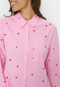 Camicia button-up rosa chiaro con collo e piccole fantasie a forma di cuore rosso. Presenta una singola tasca sul petto e un tessuto morbido.