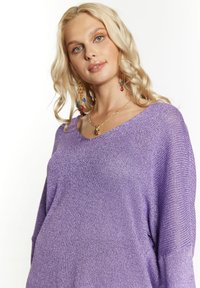 Violetinė megztinė su V-formos iškirpte, laisvu siluetu ir subtiliu metaliniu blizgesiu. Textūruota medžiaga su ilgomis rankovėmis ir plačiu raukinėtu apvadu.