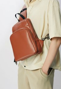 Sac en bandoulière en cuir marron avec plusieurs compartiments zippés, des sangles ajustables et un design élégant ; porté sur une chemise beige clair.
