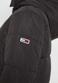 Černý prošívaný rukáv bundy s malou obdélníkovou nášivkou v b�ílé, červené a tmavě modré barvě našitou poblíž ramene.