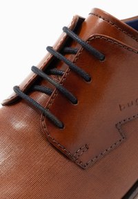 Chaussure en cuir marron avec une surface texturée, dotée de lacets noirs et de coutures détaillées. Le design comprend un panneau en tissu contrastant.