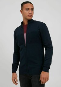 Veste zippée bleu marine en matériau lisse avec un panneau texturé sur la poitrine. Dispose de manches longues et de poignets et d'un ourlet côtelés. Stylisée avec un intérieur bordeaux.