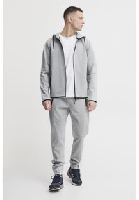 Light gray athletic tracksuit with a zip-up hoodie, white t-shirt underneath, and tapered pants. Sneakers are dark with contrasting accents.