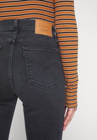 Jeans in denim scuro con vita alta, caratterizzati da due tasche posteriori e un'etichetta in pelle marrone. Maglia a maniche lunghe a righe parzialmente visibile.