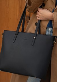 Zwarte totebag met lange, platte handvatten, gemaakt van een gestructureerd materiaal. Beschikt over gouden hardware en een subtiele logo op de voorkant.