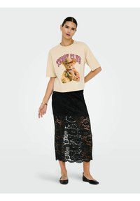 Kreemjas lühikeste varrukatega t-särk karu kujutise ja tekstiga "TEDDY CLUB", kombineeritud musta pitsilise lillemustriga midi seelikuga.