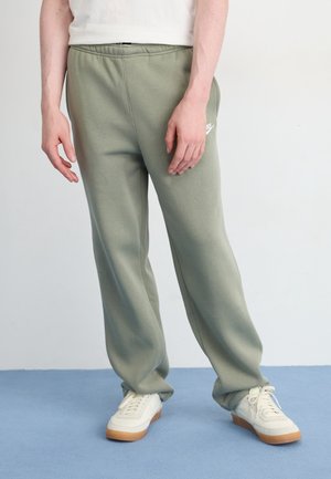 Person trägt hellgrüne Nike-Sweatpants, weiße Sneakers mit Gummisohlen und ein weißes Hemd vor einem schlichten Hintergrund.