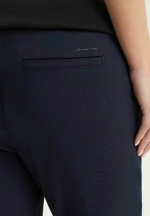 Zbliżenie osoby noszącej granatowe spodnie z tylną wszywaną kieszenią i logo "diverse", ręka spoczywa wzdłuż prawego uda.