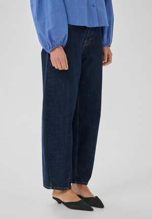 Personne portant un jean bleu foncé à jambes larges, une chemise bleue à manches longues et des chaussures noires à bout pointu et à talons bas, debout sur un fond uni.