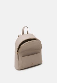 Beige reppu, joka on valmistettu sileästä kankaasta ja sen muoto on pyöreä. Repussa on suuri etupuolen vetoketjullinen tasku ja kultainen vetoketju koristeena.