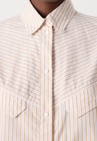 Gros plan sur une personne portant une chemise beige et blanche à rayures verticales, à boutons, avec un col pointu et deux poches poitrine.