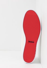 Suela de zapatilla con patrón de goma roja, entresuela blanca y parte superior beige, con un logo negro en el lateral.
