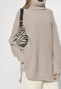 Femme portant un pull à col roulé beige tricoté avec des fentes sur les côtés, un pantalon blanc cassé, et un sac d'épaule imprimé zèbre avec une sangle noire.