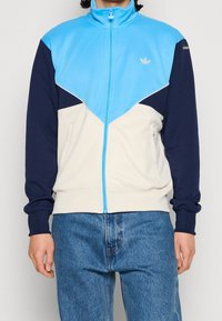 Träningsjacka med en ljusblå överdel, marinblå ärmar och krämfärgad nederdel. Dragkedja, ribbade muddar och broderad logotypdetalj.