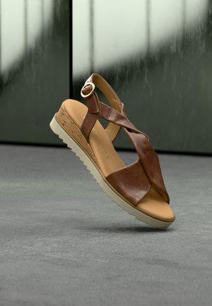 Sandale en cuir marron avec des lanières croisées, une semelle compensée recouverte de liège, et une boucle métallique ronde en guise d'accent. Semelle texturée de couleur claire.