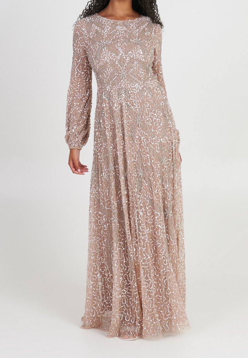Femme portant une robe longue nude à manches longues avec des motifs complexes en sequins argentés et une taille ajustée.