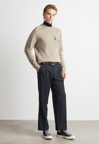 Camicia beige a maniche lunghe con tasca, abbinata a pantaloni neri plissettati e sneakers nere. Texture liscia, vestibilità casual e design moderno.