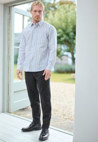 Gestreifte Button-Down-Hemd in Hellblau und Weiß, kombiniert mit dunkel gefitteten Hosen und schwarzen Leder-Dressschuhen; Hände an den Seiten.