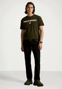 Young man standing wearing olive green "Polo Sport" t-shirt, black pants, black watch, and dark green sneakers against a plain white background.