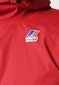 Veste rouge avec un logo K-Way bleu, blanc et rouge sur la poitrine et un cordon noir à la capuche.