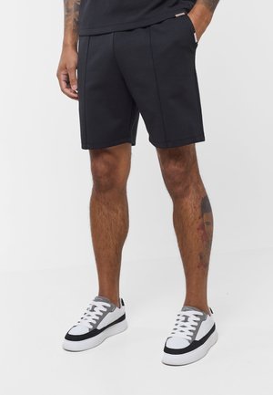 Άνδρας με μαύρα σορτς μέχρι το γόνατο, μαύρα και λευκά αθλητικά παπούτσια, με το ένα χέρι στην τσέπη, στέκεται μπροστά σε απλό φόντο.