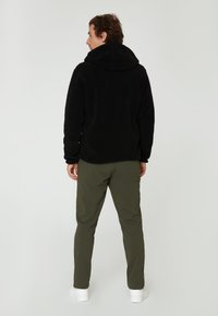 Zwarte fleece hoodie met een grote capuchon; gedragen met olijfgroene broek en witte schoenen. Weergave van achteraf, met textuurstof.