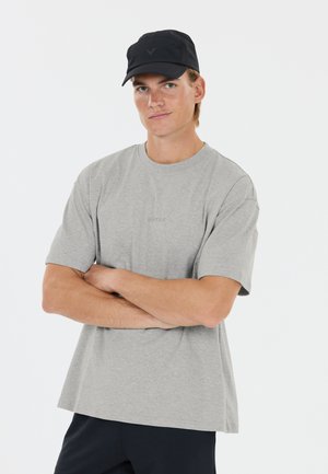 Junger Mann trägt ein hellgraues, locker sitzendes T-Shirt und eine schwarze Kappe, steht mit verschränkten Armen und schaut mit einem leichten Lächeln nach vorne.