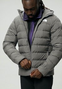 Jachetă puffer gri cu închidere cu fermoar, manșete bătute și glugă. Prezintă un design matlasat și logo-ul The North Face pe umărul stâng.